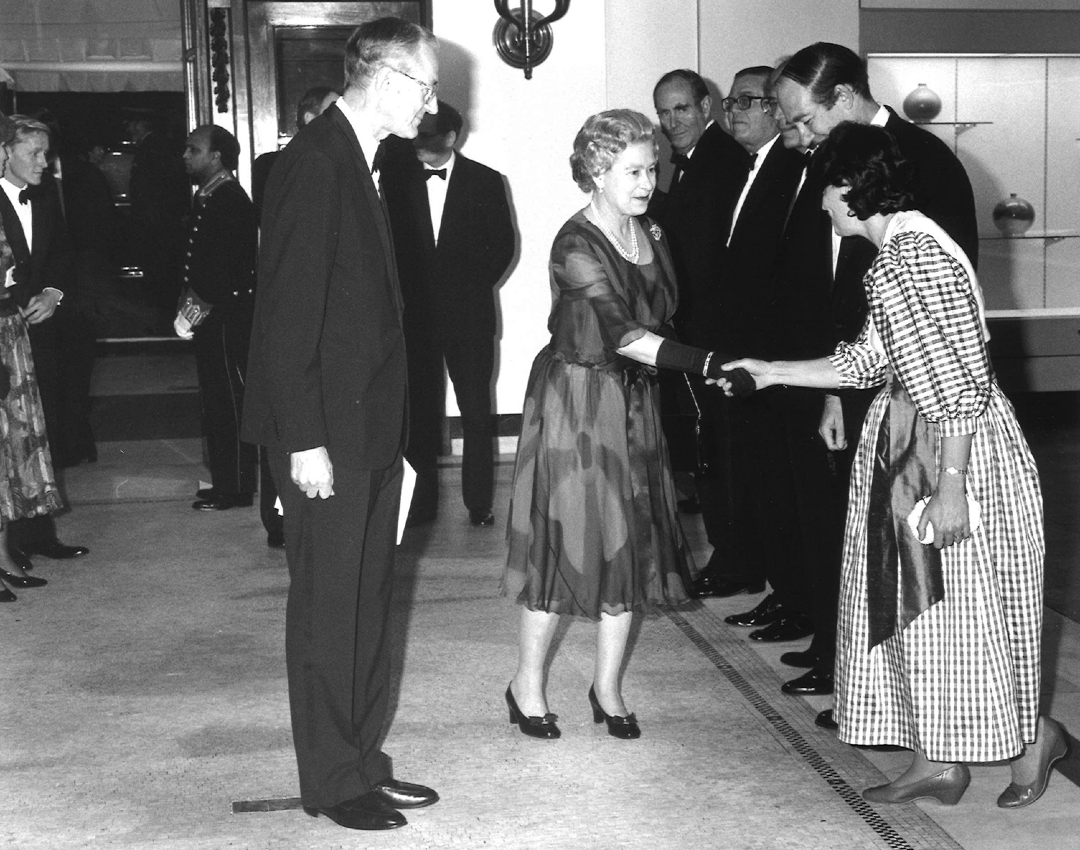 Une photo en noir et blanc de la reine, escortée par Lord Windlesham, président des administrateurs du British Museum, rencontrant des dignitaires et serrant la main d'une femme en robe à carreaux. 