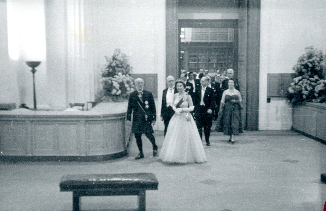 Une photographie en noir et blanc de la reine portant une robe longue, un diadème et une ceinture, et entrant dans une pièce.  