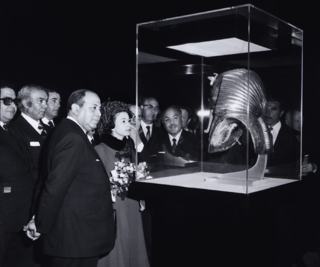 Une photographie en noir et blanc de la Reine regardant une vitrine contenant le masque mortuaire de Toutankhamon. 