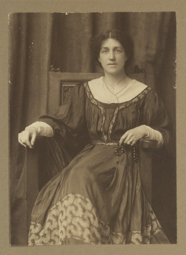 Photo sépia de May Morris assise sur une chaise en robe, les bras reposant sur les accoudoirs de la chaise et regardant droit dans la caméra.